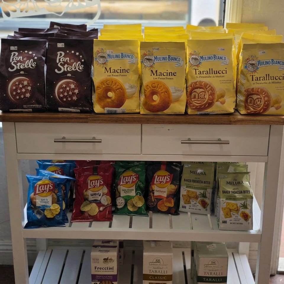 Display cabinet stocked with Italian biscuits, potato chips, savory snacks, and packaged taralli beside a restaurant window.