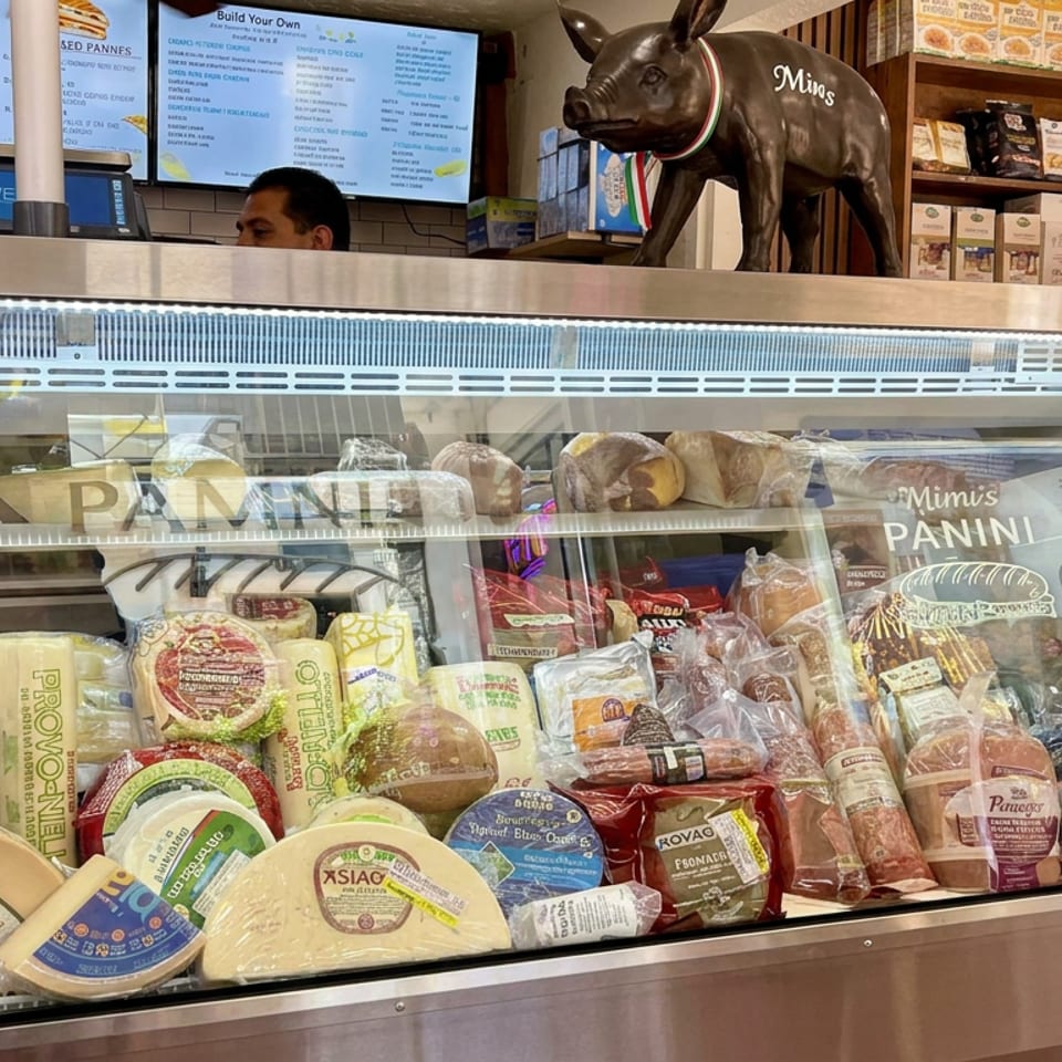 Mimi’s Panini deli counter displaying assorted cheeses, cured meats, bread, and sandwiches beneath menu boards and a pig statue.